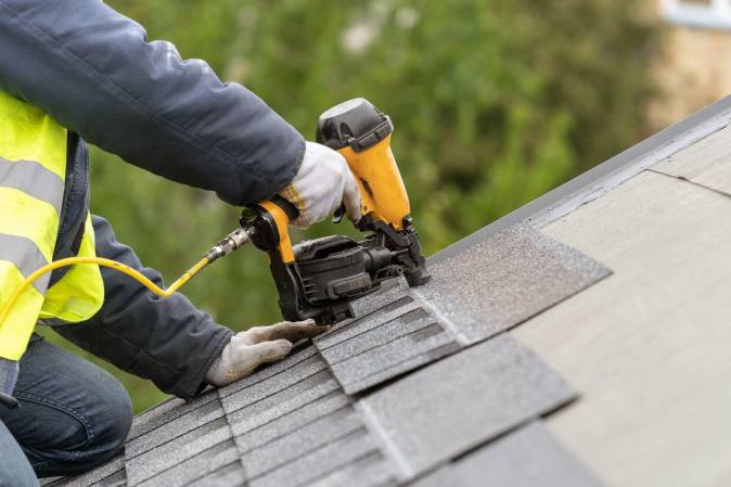 worker drilling shingles on a house rooftop